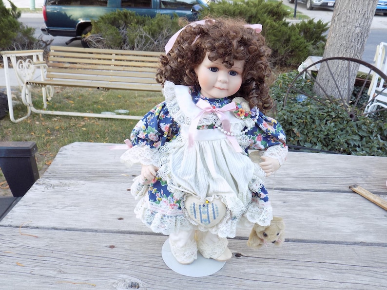 May include: A porcelain doll with brown curly hair, wearing a blue floral dress with white lace trim and a white apron. The doll has a sweet expression and is holding a small stuffed animal. A heart-shaped patch is sewn on the apron.