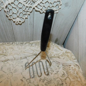 May include: A vintage potato masher with a black handle and a stainless steel mashing head. The handle has a hole for hanging. The masher is designed for kitchen use and is shown against a lace tablecloth.
