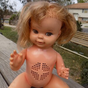 May include: A vintage plastic doll with blonde hair and brown eyes. The doll has a peach-colored body with a square pattern on the chest. It is sitting on a wooden surface outdoors.