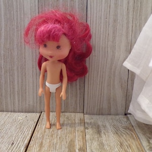 Pode incluir: Uma pequena boneca de pl&aacute;stico com cabelo rosa e uma roupa interior branca. A boneca est&aacute; em p&eacute; sobre uma superf&iacute;cie de madeira.