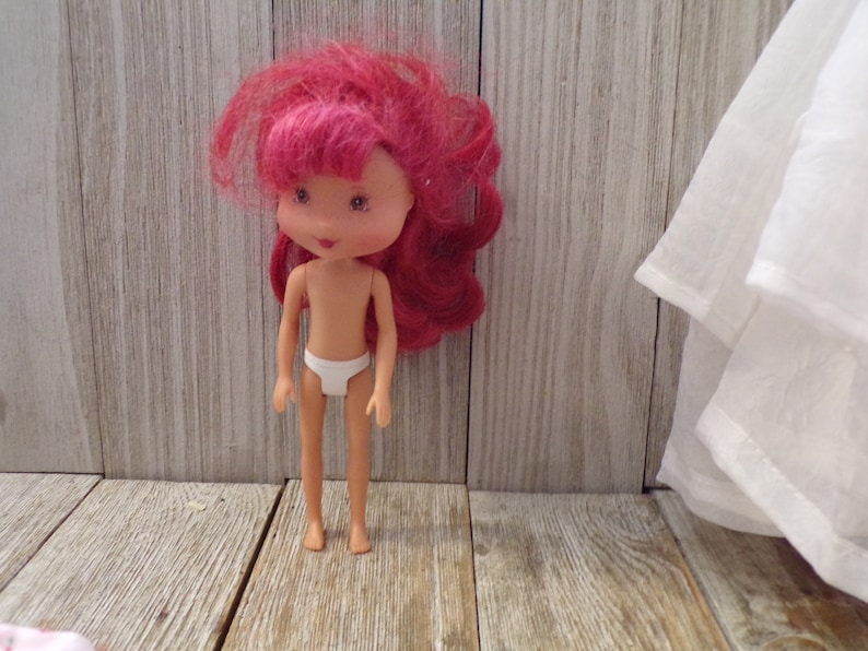 Pode incluir: Uma pequena boneca de pl&aacute;stico com cabelo rosa e uma roupa interior branca. A boneca est&aacute; em p&eacute; sobre uma superf&iacute;cie de madeira.