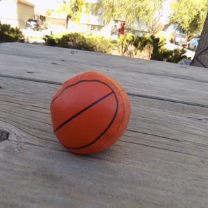 May include: An orange basketball stress ball with black lines, resting on a weathered wooden surface. The ball's design mimics a real basketball, offering a tactile and playful experience. The background shows a natural outdoor setting.