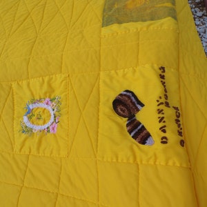May include: A bright yellow quilt with a quilted diamond pattern. The quilt features embroidered patches with floral and abstract designs. One patch has the name "Danny" embroidered on it.