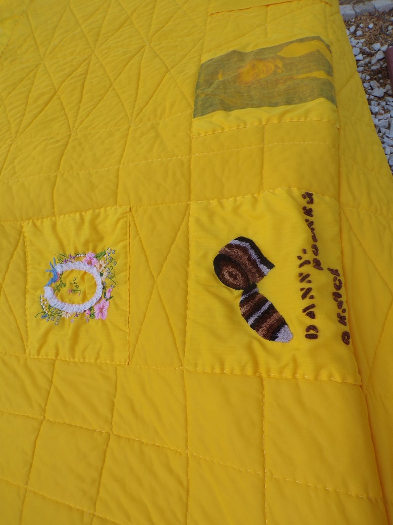 May include: A bright yellow quilt with a quilted diamond pattern. The quilt features embroidered patches with floral and abstract designs. One patch has the name "Danny" embroidered on it.