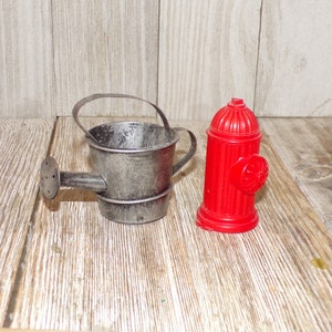 May include: A small, silver watering can and a red plastic fire hydrant. Both are miniature in size and appear to be decorative.