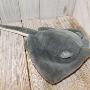 May include: A gray plush stingray toy with a white underside. The toy is lying on a wooden surface.