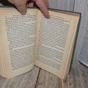 May include: A close-up of an open book with green cover and cream-colored pages. The text on the page reads "The Enormous Room" and "Begin a Pilgrimage".