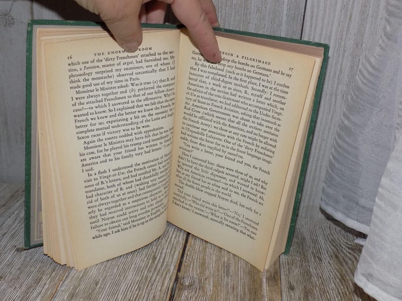 May include: A close-up of an open book with green cover and cream-colored pages. The text on the page reads "The Enormous Room" and "Begin a Pilgrimage".