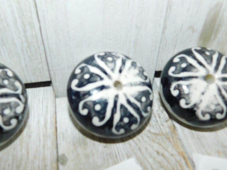 K&ouml;nnte beinhalten: Drei Keramik-Kn&ouml;pfe mit einem wei&szlig;en und schwarzen Blumendesign. Die Kn&ouml;pfe sind rund und haben ein Loch in der Mitte.