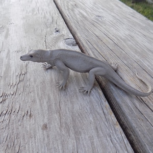 May include: A gray lizard figurine on a wooden surface. The lizard is posed in a walking position with its tail curled.