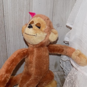 May include: A plush monkey toy with brown fur, a light tan face, and a small pink bow. The monkey has closed eyes and a stitched smile. Its arms are outstretched, and it is sitting down.