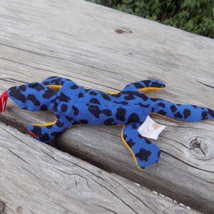 May include: Blue and black spotted plush toy lizard with yellow accents and a red tongue. The lizard is lying on a wooden surface.
