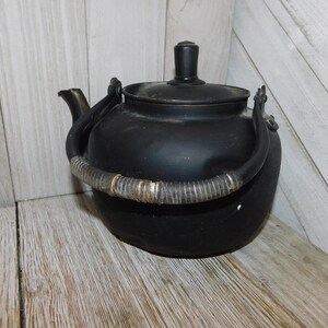 May include: Black metal teapot with a woven handle. The teapot has a lid and a spout. The teapot is sitting on a wooden surface.