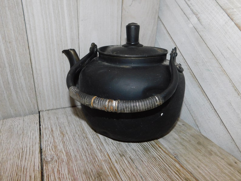 May include: Black metal teapot with a woven handle. The teapot has a lid and a spout. The teapot is sitting on a wooden surface.