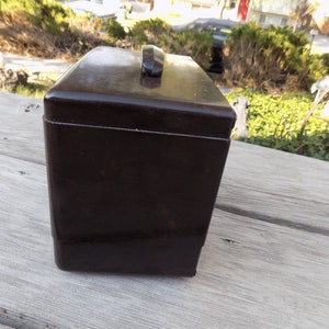 Puede incluir: Una caja rectangular marr&oacute;n oscuro con un asa en la parte superior. La caja tiene un acabado brillante y parece estar hecha de un material s&oacute;lido. El asa es de un tono marr&oacute;n m&aacute;s oscuro. La caja est&aacute; sobre una superficie de madera.