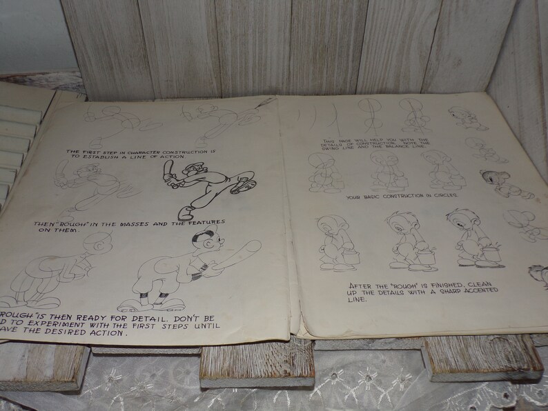 May include: Vintage open book with pencil sketches of cartoon character construction. The pages show step-by-step instructions, including establishing a line of action and roughing in masses and features. The text includes instructions on how to draw the characters.