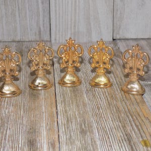 May include: Five gold-coloured metal place card holders with ornate designs. Each holder has a round base and a decorative top.