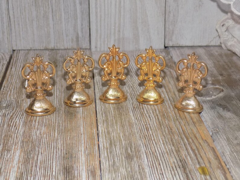May include: Five gold-coloured metal place card holders with ornate designs. Each holder has a round base and a decorative top.
