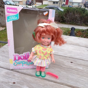 May include: A red-haired doll with a watermelon print dress and green shoes. The doll is holding a pink comb. The doll is in a box that says "Playskool Polly Surprise Doll Surprise".