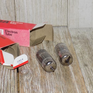 May include: Two vintage RCA Electron Tube boxes and two glass vacuum tubes on a wooden surface. The red boxes have white text and the words "Electron Tube". The tubes are cylindrical with metal bases.