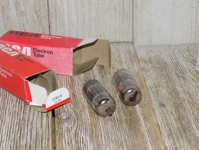 May include: Two vintage RCA Electron Tube boxes and two glass vacuum tubes on a wooden surface. The red boxes have white text and the words "Electron Tube". The tubes are cylindrical with metal bases.