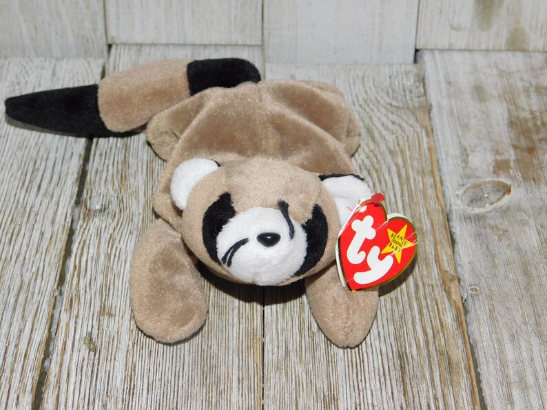 K&ouml;nnte beinhalten: Ein braunes und schwarzes Waschb&auml;r-Beanie Baby mit wei&szlig;em Gesicht und schwarzen Augen. Das Beanie Baby liegt auf dem Bauch mit dem Schwanz hinter sich eingerollt. Das Beanie Baby hat ein rotes und wei&szlig;es Ty-Etikett, das "Beanie Babies" sagt und einen Stern darauf hat.