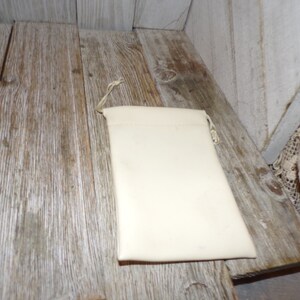 Puede incluir: Una bolsa blanca de cord&oacute;n con un dise&ntilde;o sencillo, hecha de una tela suave. La bolsa est&aacute; sobre una superficie de madera.
