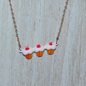 May include: A gold chain necklace with three cupcake charms. The cupcakes are white with pink frosting and orange bottoms.