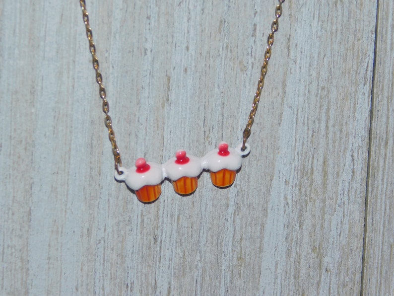 May include: A gold chain necklace with three cupcake charms. The cupcakes are white with pink frosting and orange bottoms.