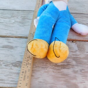 May include: A blue plush toy with yellow feet and a white face. The toy is lying on a wooden surface.