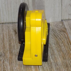 May include: A yellow and black vintage toy, possibly a rotary phone or a similar device. The toy has a black circular component and a yellow rectangular body. It sits on a black base.