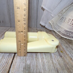May include: A pair of vintage off-white plastic hair curlers, measuring approximately 7 inches in length, are displayed on a wooden surface. The curlers are designed with a rounded shape and a textured surface.