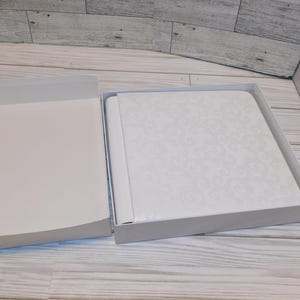 May include: An open white box containing a white album with a floral pattern. The album is inside a white box. The box is on a light wood surface.