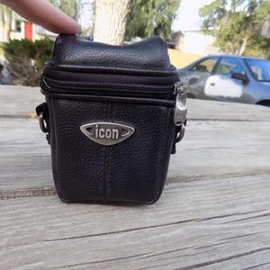 May include: A black textured camera bag with a zippered top closure and a silver-toned metal emblem on the front featuring the word icon. The bag has a structured rectangular shape with rounded corners and metal hardware attachments on the sides.