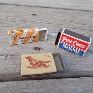 May include: Three vintage matchboxes are displayed on a weathered wooden surface. One box is orange and white with "hungry tiger" text. Another is red, white, and blue with "Fire Chief Matches" text. The third box features a dog illustration.