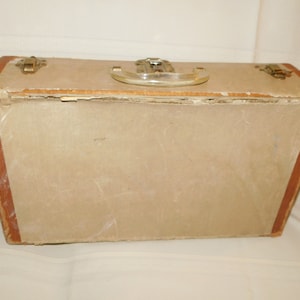 May include: A rectangular suitcase with a light beige center panel and textured brown edges. The top features a clear plastic handle and two metallic latches on either side. The edges show signs of wear and fraying along the seams of the case.