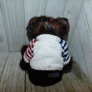 May include: A brown teddy bear wearing a white knitted jumper with red and blue stripes. The jumper has a crew neck and the bear is sitting on a wooden surface.