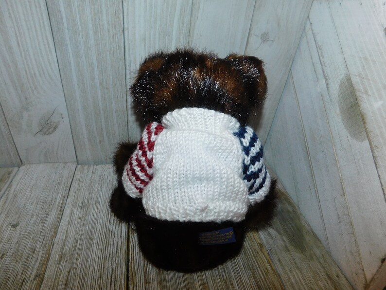 May include: A brown teddy bear wearing a white knitted jumper with red and blue stripes. The jumper has a crew neck and the bear is sitting on a wooden surface.