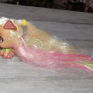 May include: A tan puppy figurine with blonde hair and pink and green streaks. The puppy has brown ears, a green collar, and pink paw pads. The hair is styled with a yellow and pink hair tie.
