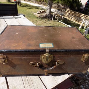 May include: Vintage brown trunk with brass hardware, black trim, and a leather handle. The trunk has corner protectors and a central clasp. A small label is affixed to the top. The trunk is sitting on a wooden surface.