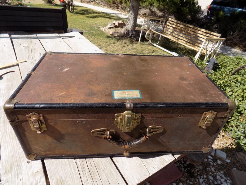 May include: Vintage brown trunk with brass hardware, black trim, and a leather handle. The trunk has corner protectors and a central clasp. A small label is affixed to the top. The trunk is sitting on a wooden surface.