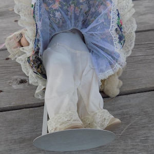 May include: A vintage doll with white lace-trimmed pants and cream-colored shoes. The doll's dress features a floral pattern with blue and pink accents, and a white lace trim. The doll is mounted on a metal stand.