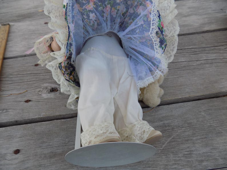 May include: A vintage doll with white lace-trimmed pants and cream-colored shoes. The doll's dress features a floral pattern with blue and pink accents, and a white lace trim. The doll is mounted on a metal stand.