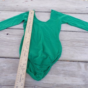 Puede incluir: Un leotardo de manga larga de color verde brillante con cuello redondo. El leotardo est&aacute; colocado plano sobre una superficie de madera con una regla al lado.