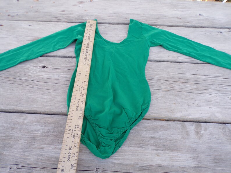 Puede incluir: Un leotardo de manga larga de color verde brillante con cuello redondo. El leotardo est&aacute; colocado plano sobre una superficie de madera con una regla al lado.