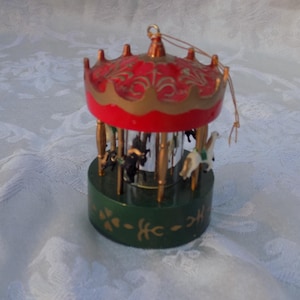 May include: A carousel ornament featuring a red roof with gold decorative accents, a dark green cylindrical base with gold floral patterns, and small horse figures positioned around a central mirrored column with a gold hanging loop at the top.