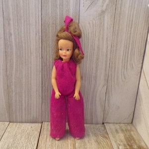 May include: A vintage doll with brown hair and a pink jumpsuit. The doll has a bow in her hair and is standing on a wooden surface.
