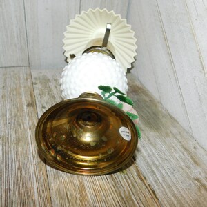 May include: A white milk glass lamp with a brass base and a white paper shade. The lamp has a floral design with green leaves and pink flowers.