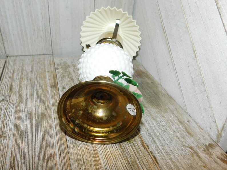May include: A white milk glass lamp with a brass base and a white paper shade. The lamp has a floral design with green leaves and pink flowers.
