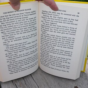 May include: Open book with yellow pages, showing a scene from a Nancy Drew mystery novel. The text reads "The Secret of Shadow Ranch" and "The men may come back. Let's wait here and see."